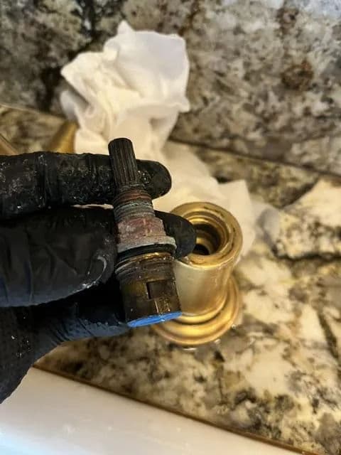 Old faucet cartridge held in a gloved hand, with a gold fixture and cleaning cloth in background.