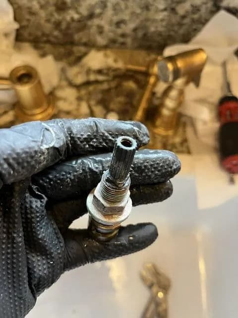 Hand holding a worn spark plug with tools and plumbing fixtures in the background.