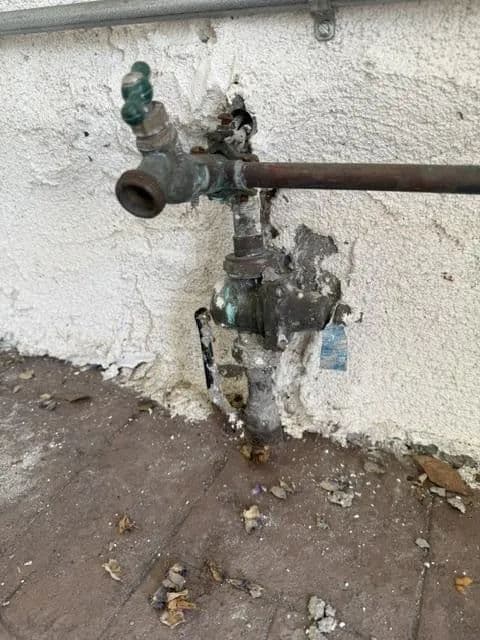 Damaged outdoor water pipe with corrosion and surrounding debris on a wall.